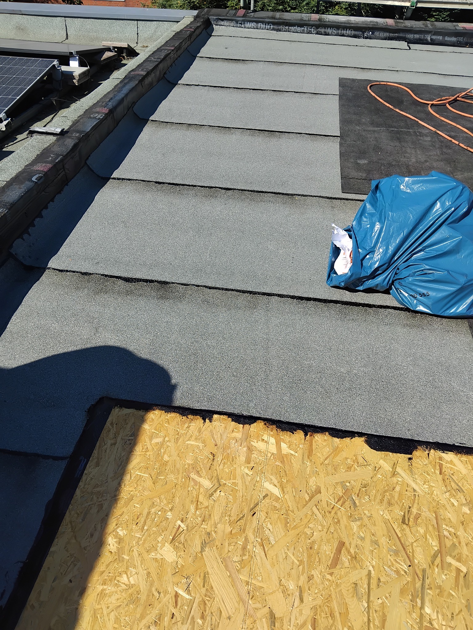 Dach pokryty papą bitumiczną, fragment płyty OSB, niebieski worek z odpadami i zwój kabla na dachu. Widoczne panele słoneczne w tle.