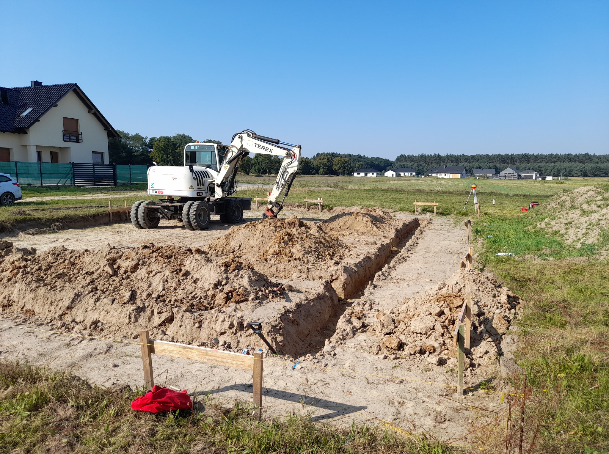 Biała koparka Terex wykonuje wykop rowu pod fundamenty na piaszczystym terenie. W tle dom jednorodzinny i łąka pod błękitnym niebem.