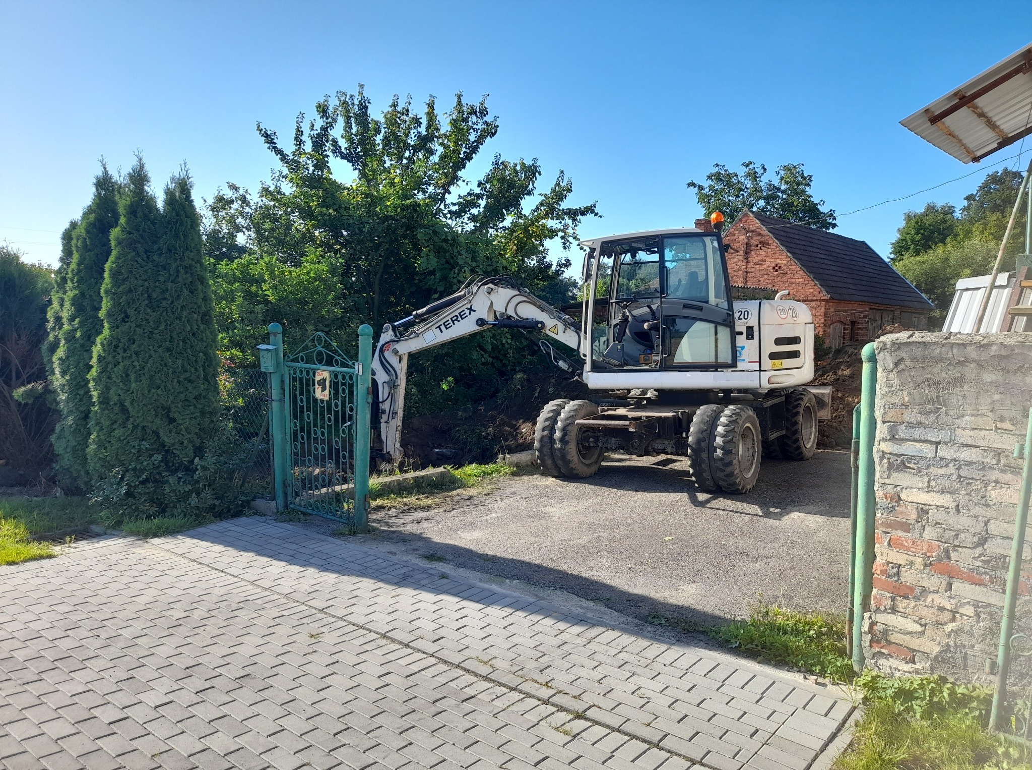 Biała koparka kołowa Terex na posesji z zielonymi bramami i kostką brukową, w tle ceglany budynek i drzewa, błękitne niebo. Maszyna gotowa do pracy.