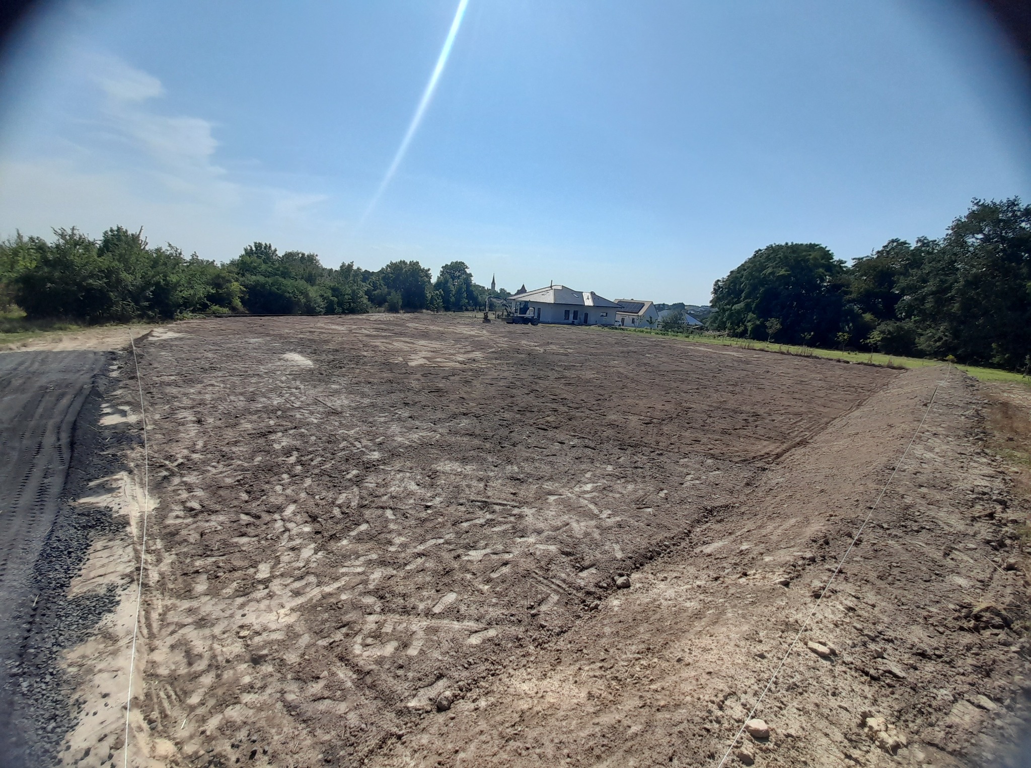 Wypoziomowany teren działki pod budowę domu, widoczne ślady opon i wyrównana ziemia. W tle budynki mieszkalne i drzewa, błękitne niebo.