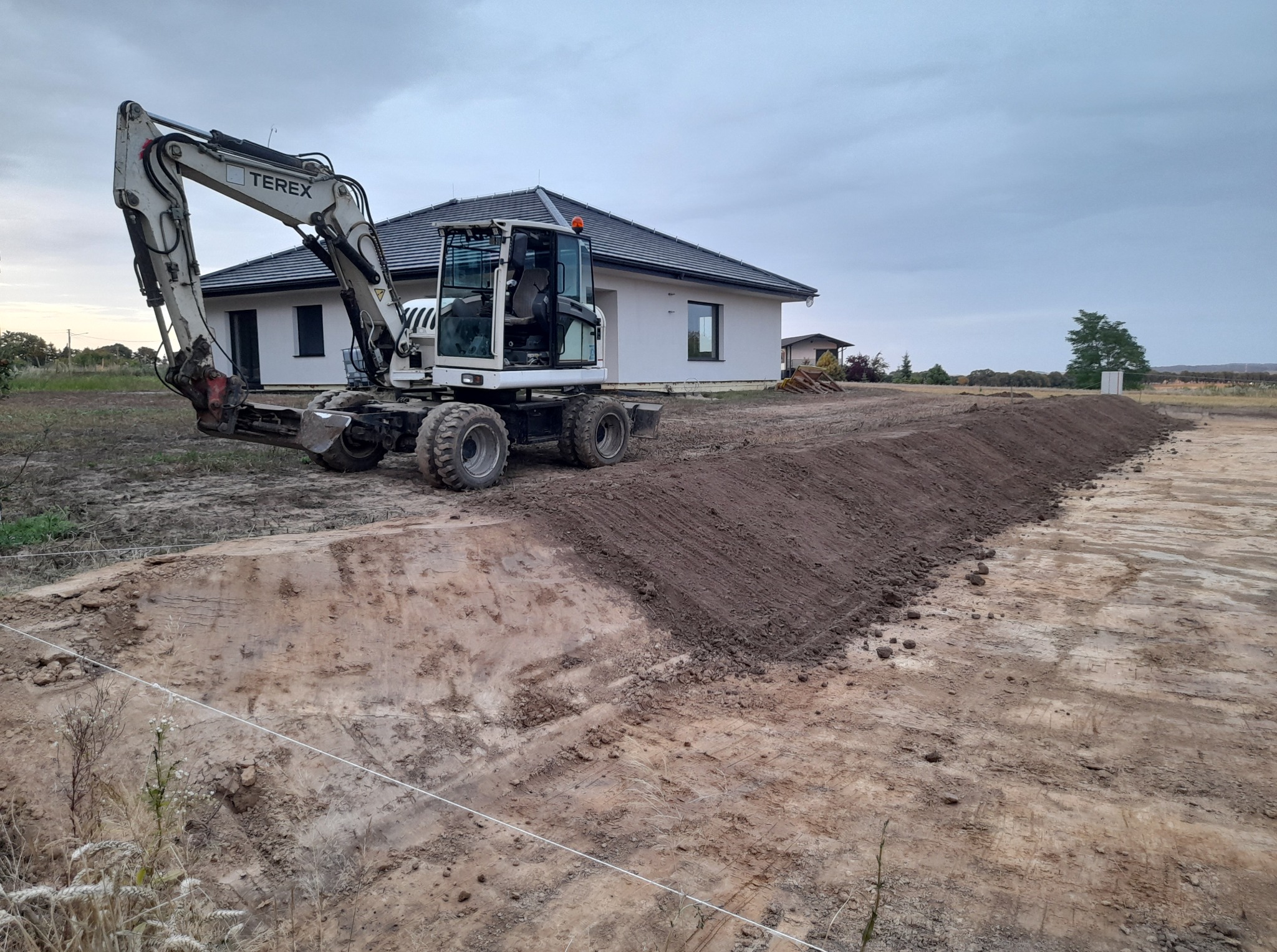 Biała koparka Terex na tle domu jednorodzinnego, widoczny wykop pod fundamenty, wstępny etap prac ziemnych.