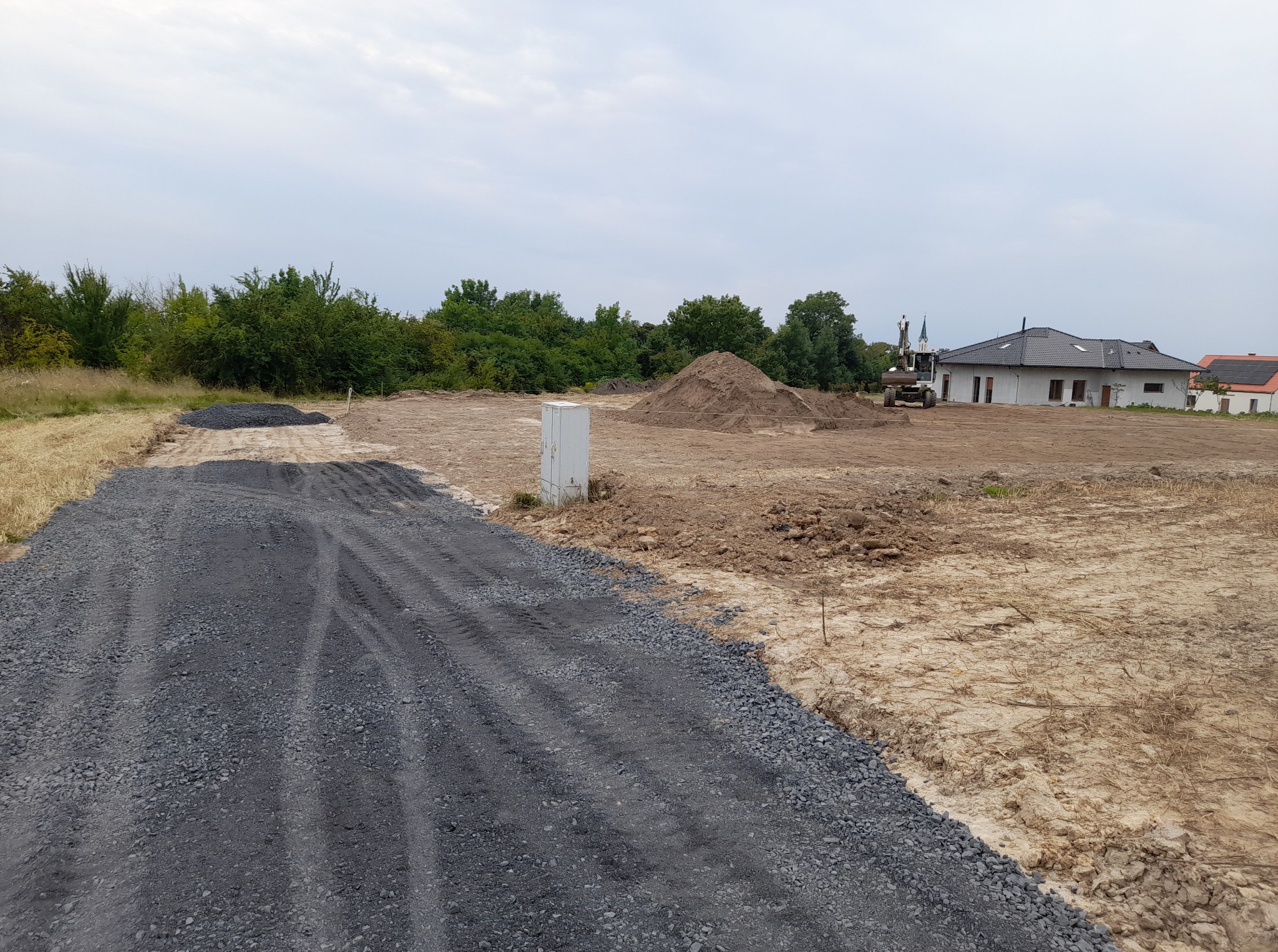 Teren budowy z utwardzoną drogą dojazdową z szarego kruszywa, w tle dom w trakcie budowy i koparka. Widok na plac z lekkiej perspektywy.