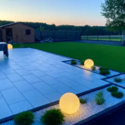 Ogród po zmroku: oświetlony taras z szarych płyt, rabaty z białym grysem, kuliste lampy, trawnik i domek narzędziowy w tle, spokojny wieczorny krajobraz.