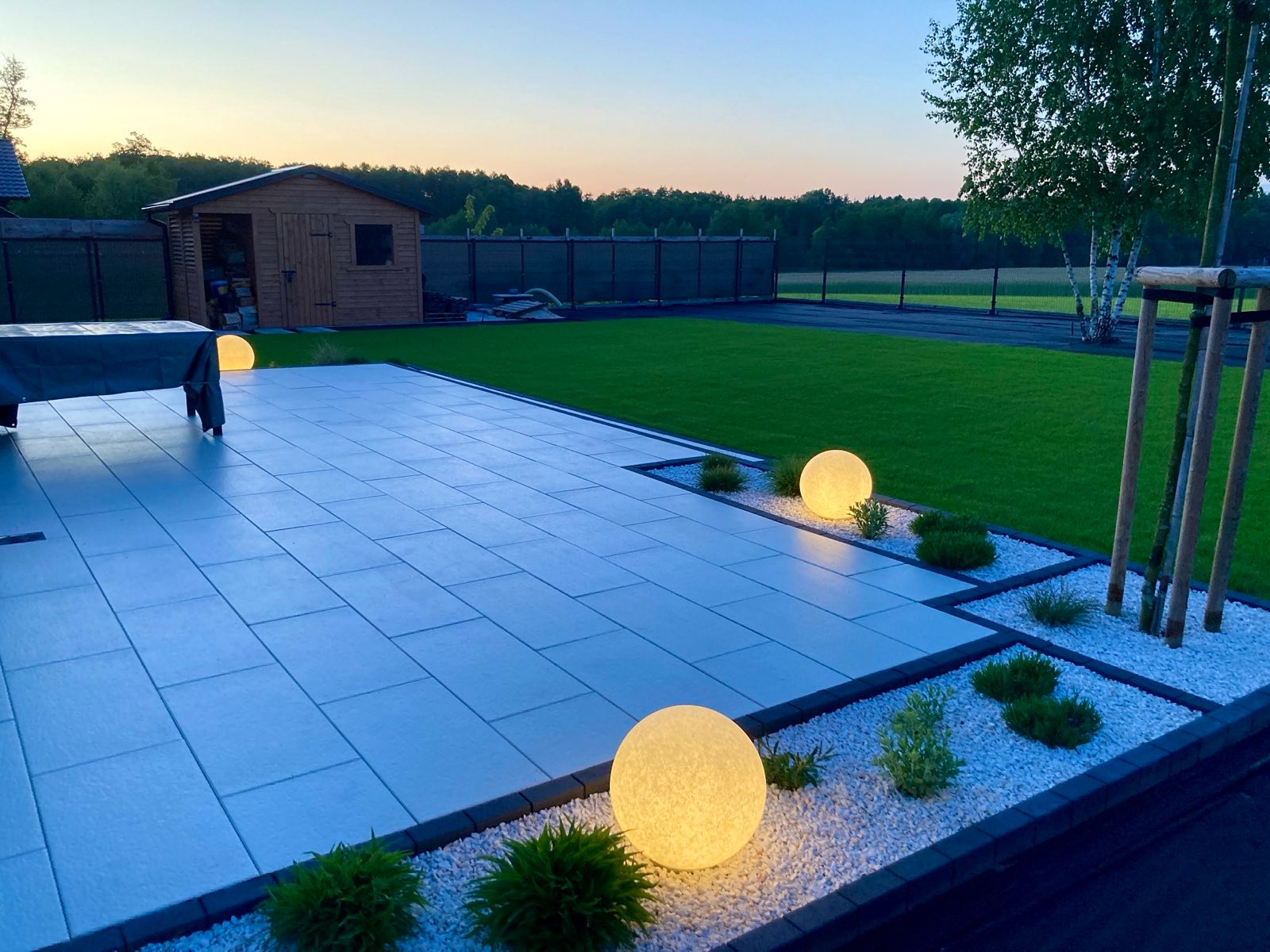 Ogród po zmroku: oświetlony taras z szarych płyt, rabaty z białym grysem, kuliste lampy, trawnik i domek narzędziowy w tle, spokojny wieczorny krajobraz.