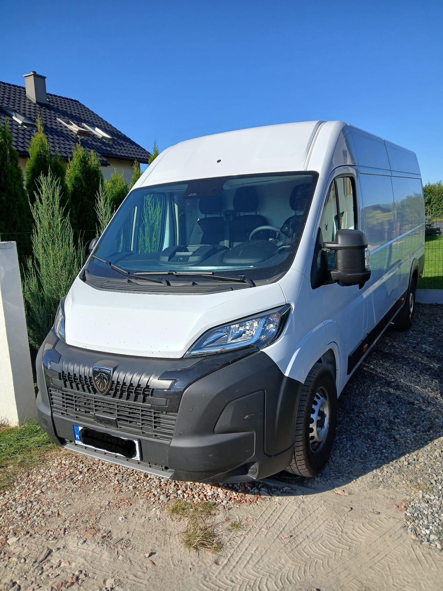 Biały Peugeot Boxer L4H2 na żwirowym podjeździe, tło: dom z ciemnym dachem i iglaki. Słoneczny dzień, błękitne niebo. Widok z przodu, lekko z boku.