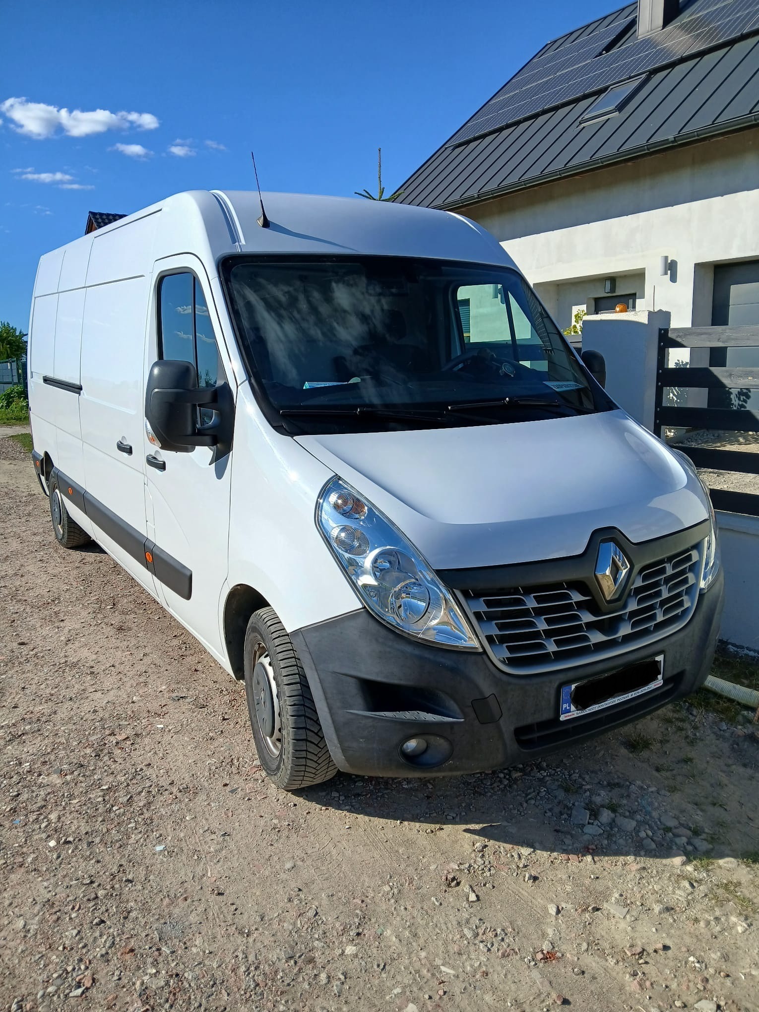 Biały bus Renault Master L3H2 na żwirowym podłożu przed nowoczesnym domem z panelami słonecznymi. Słoneczny dzień, błękitne niebo.