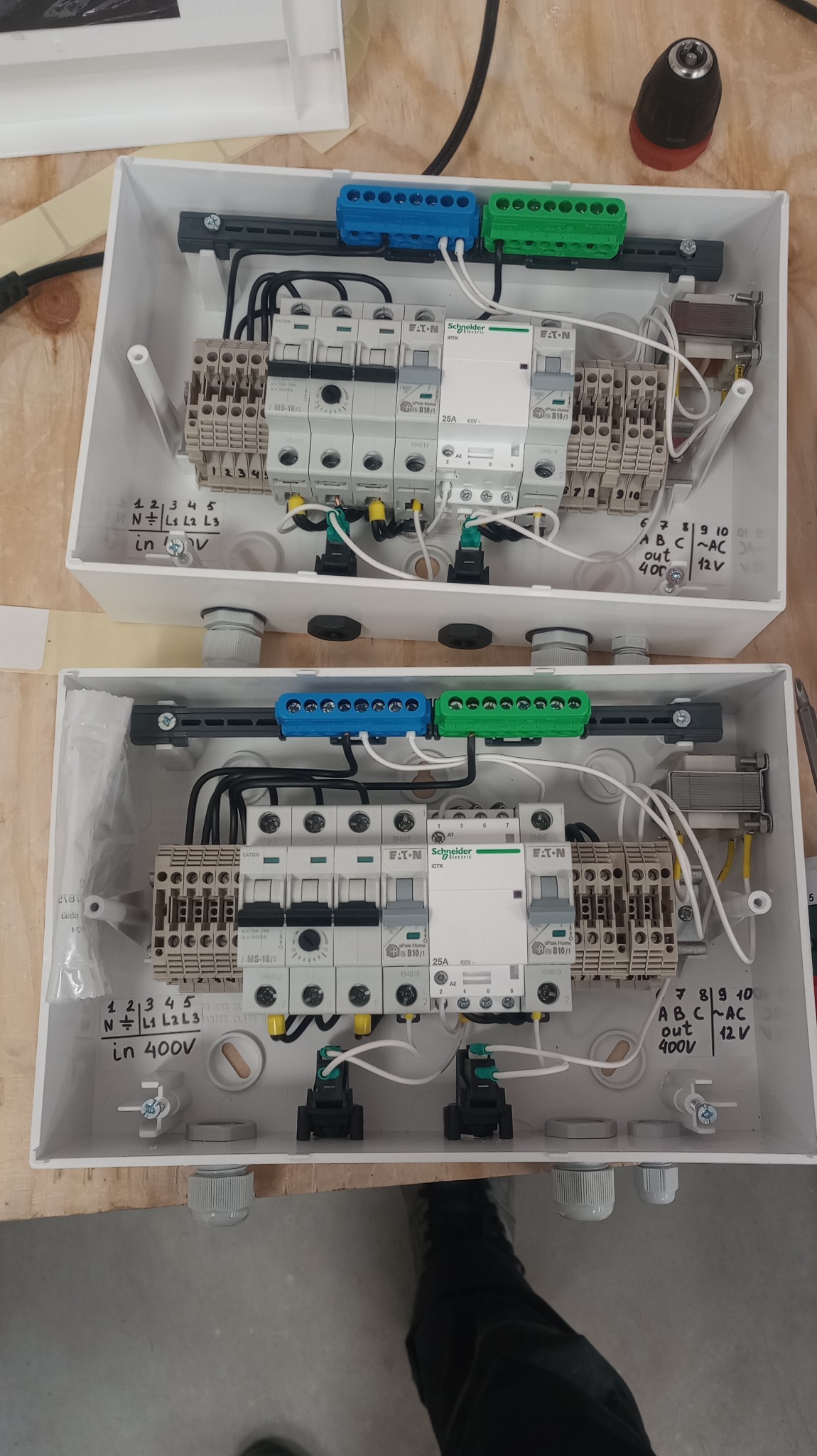 Dwa białe rozdzielacze elektryczne z zainstalowanymi bezpiecznikami, złączkami i okablowaniem na drewnianym blacie. Widoczne opisy napięć i oznaczenia.