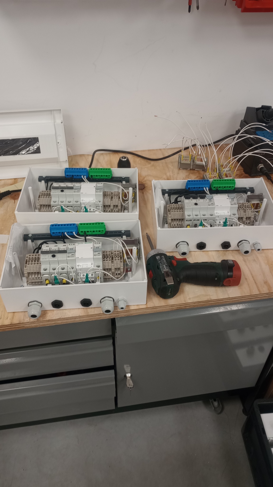 Trzy skrzynki elektryczne w trakcie montażu na warsztatowym stole, obok wkrętarka. Widoczne elementy instalacji, przewody i złączki. Detaliczne ujęcie procesu składania.