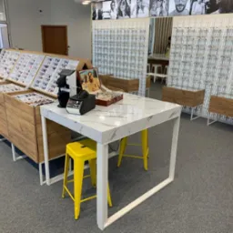 Wnętrze salonu optycznego z ekspozycją okularów na drewnianych regałach i białym stole z marmurowym blatem, żółte krzesła barowe, nowoczesny design.