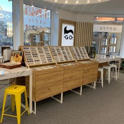 Optyk.com - Salon optyczny w Gliwicach prezentuje ekspozycję okularów na drewnianych regałach, z żółtym stołkiem i marmurowym blatem. Widoczne logo firmy i witryna z odbiciem ulicy.