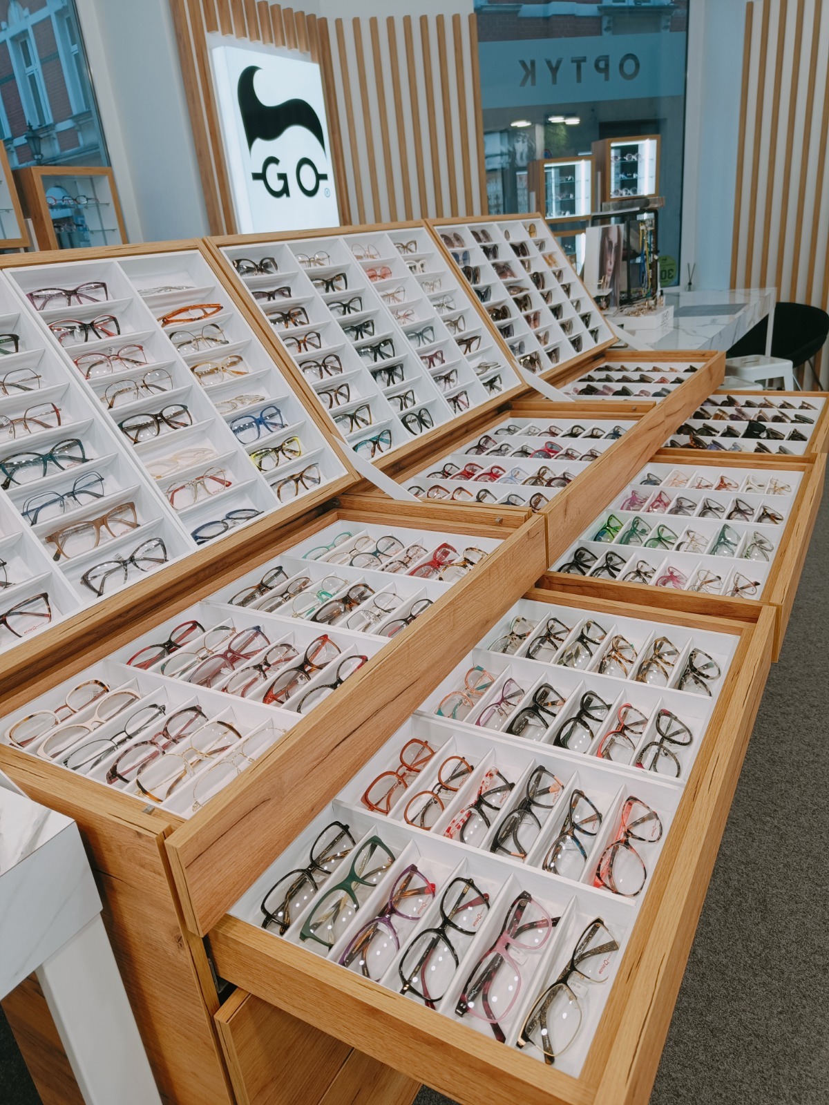 Ekspozycja różnorodnych oprawek okularów w salonie optycznym. Drewniane regały z białymi przegródkami prezentują bogaty wybór modeli.