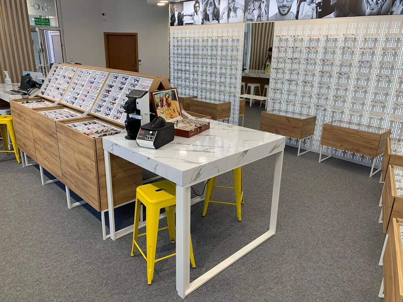 Wnętrze salonu optycznego z ekspozycją okularów na drewnianych regałach i białym stole z marmurowym blatem, żółte krzesła barowe, nowoczesny design.