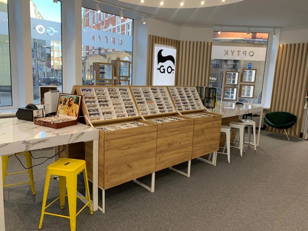 Salon optyczny w Gliwicach prezentuje ekspozycję okularów na drewnianych regałach, z żółtym stołkiem i marmurowym blatem. Widoczne logo firmy i witryna z odbiciem ulicy.