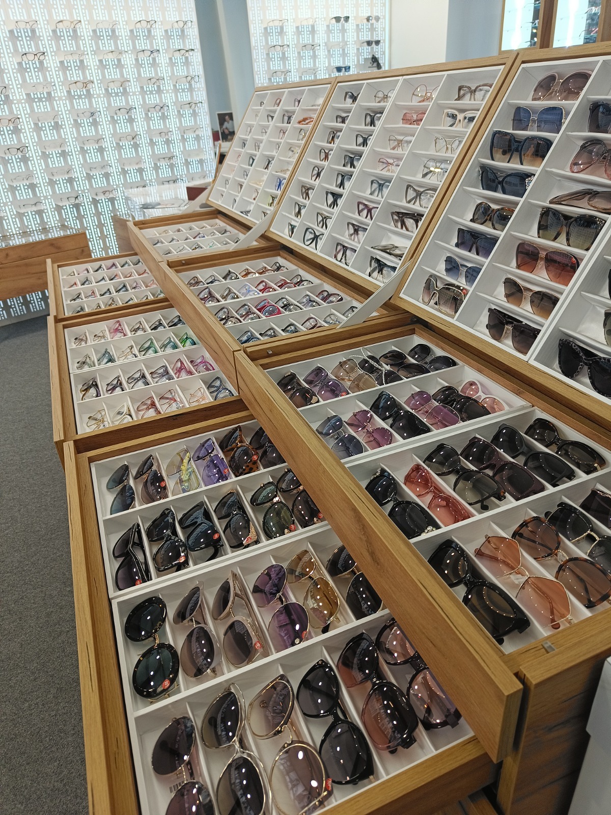 Szeroki wybór okularów korekcyjnych i przeciwsłonecznych w salonie optycznym, eksponowanych w drewnianych, białych gablotach. Różne modele i style.