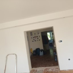 Dawid Michalak - Wnętrze po świeżym malowaniu ścian na biało, widoczne punkty oświetleniowe w suficie, drabina malarska i otwarte przejście do innego pomieszczenia.