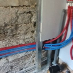 Dawid Michalak - Instalacja wodna z rurami PEX w kolorach czerwonym i niebieskim, zamontowana na ścianie z surowej cegły, obok metalowej konstrukcji. Widoczny etap prac wykończeniowych.