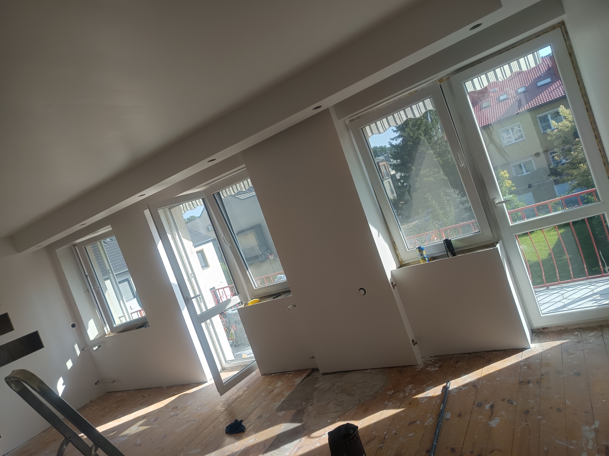 Jasne wnętrze w trakcie prac wykończeniowych: surowe ściany, okna z widokiem na zewnątrz, drewniana podłoga z resztkami farby i drabina. Etap przygotowania do malowania.