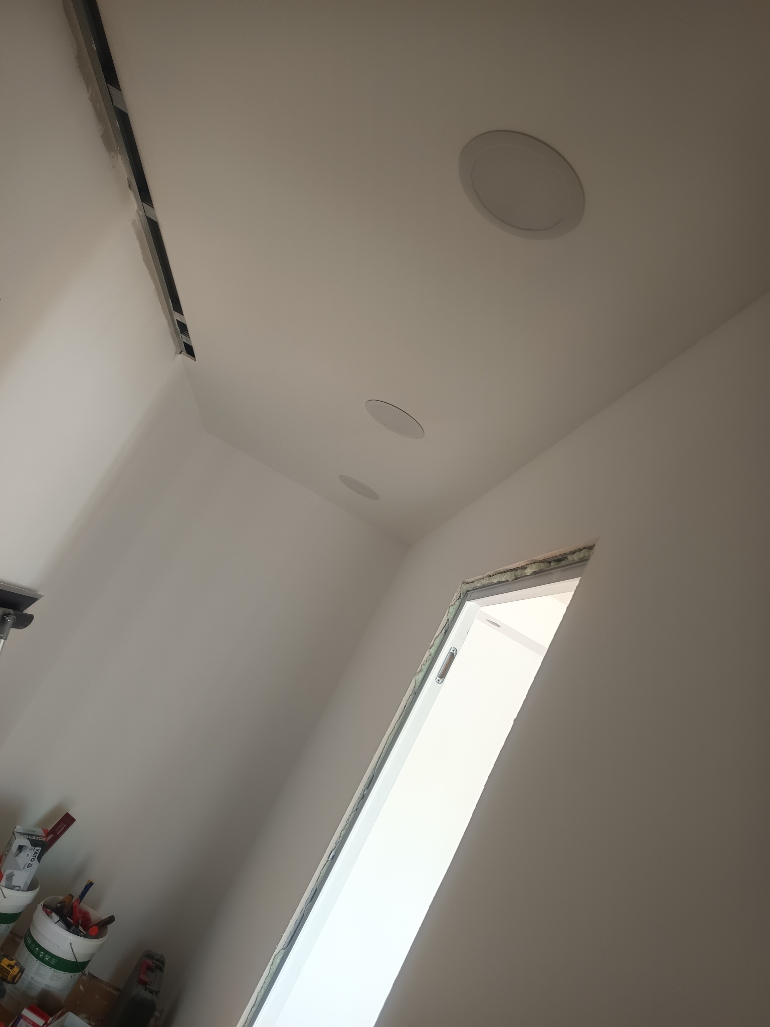 Białe wnętrze po remoncie: gładkie ściany, sufit z trzema okrągłymi lampami, ościeżnica drzwiowa z pianką montażową, puszki z farbą w rogu.