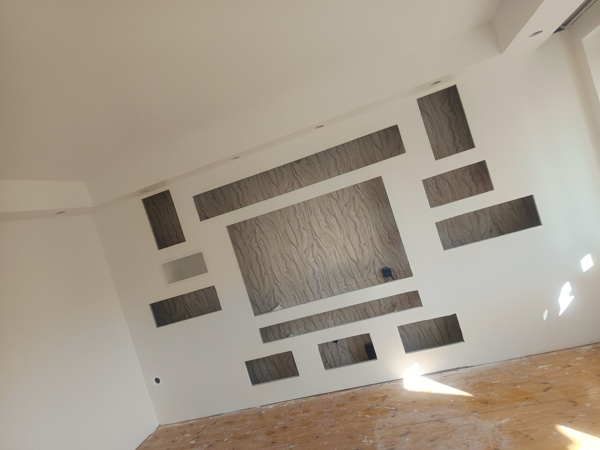 Ściana z dekoracyjnymi wnękami i panelami o marmurkowym wzorze, nowoczesne wykończenie wnętrza, jasne kolory, drewniana podłoga w trakcie prac.