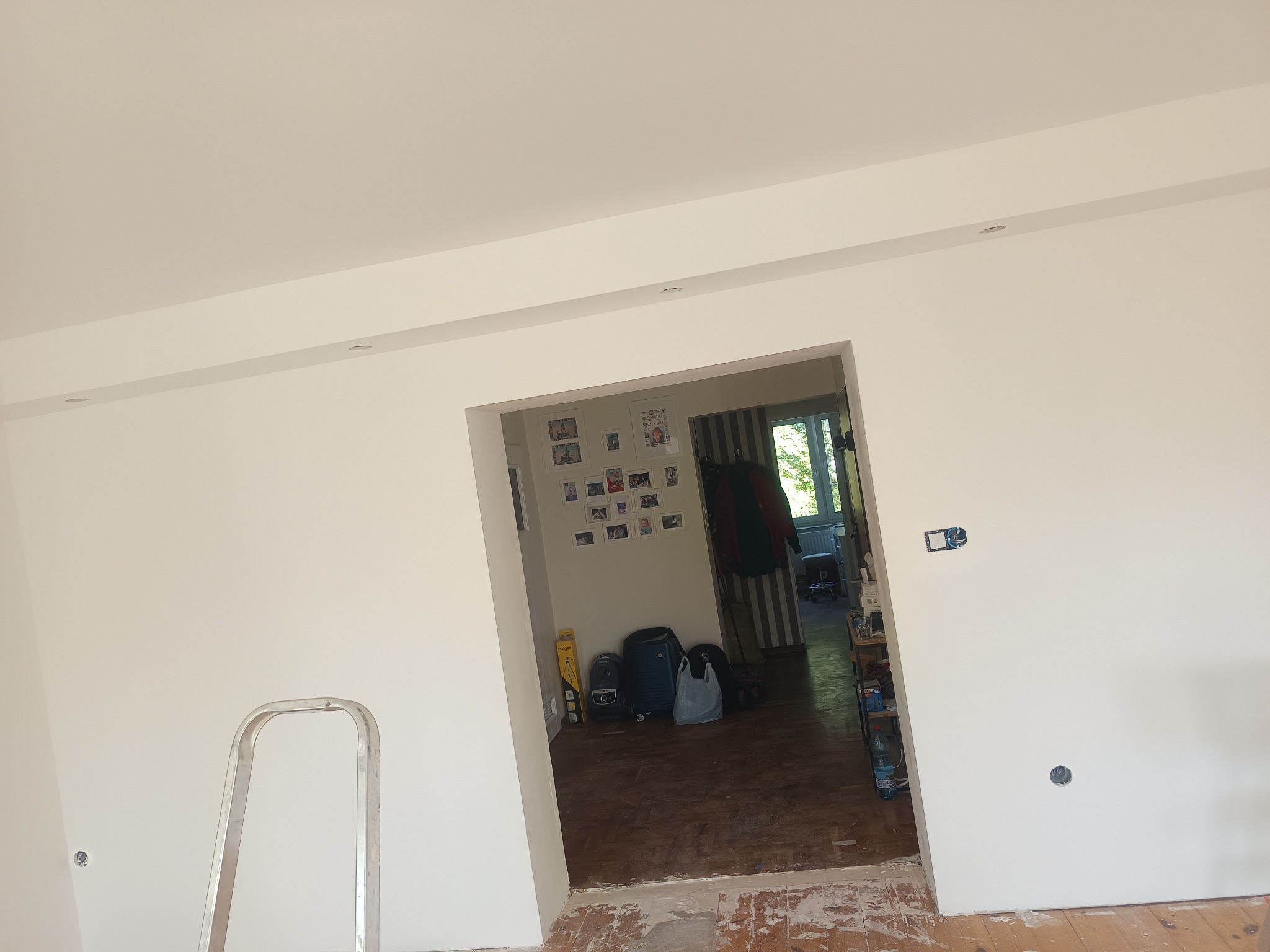 Wnętrze po świeżym malowaniu ścian na biało, widoczne punkty oświetleniowe w suficie, drabina malarska i otwarte przejście do innego pomieszczenia.