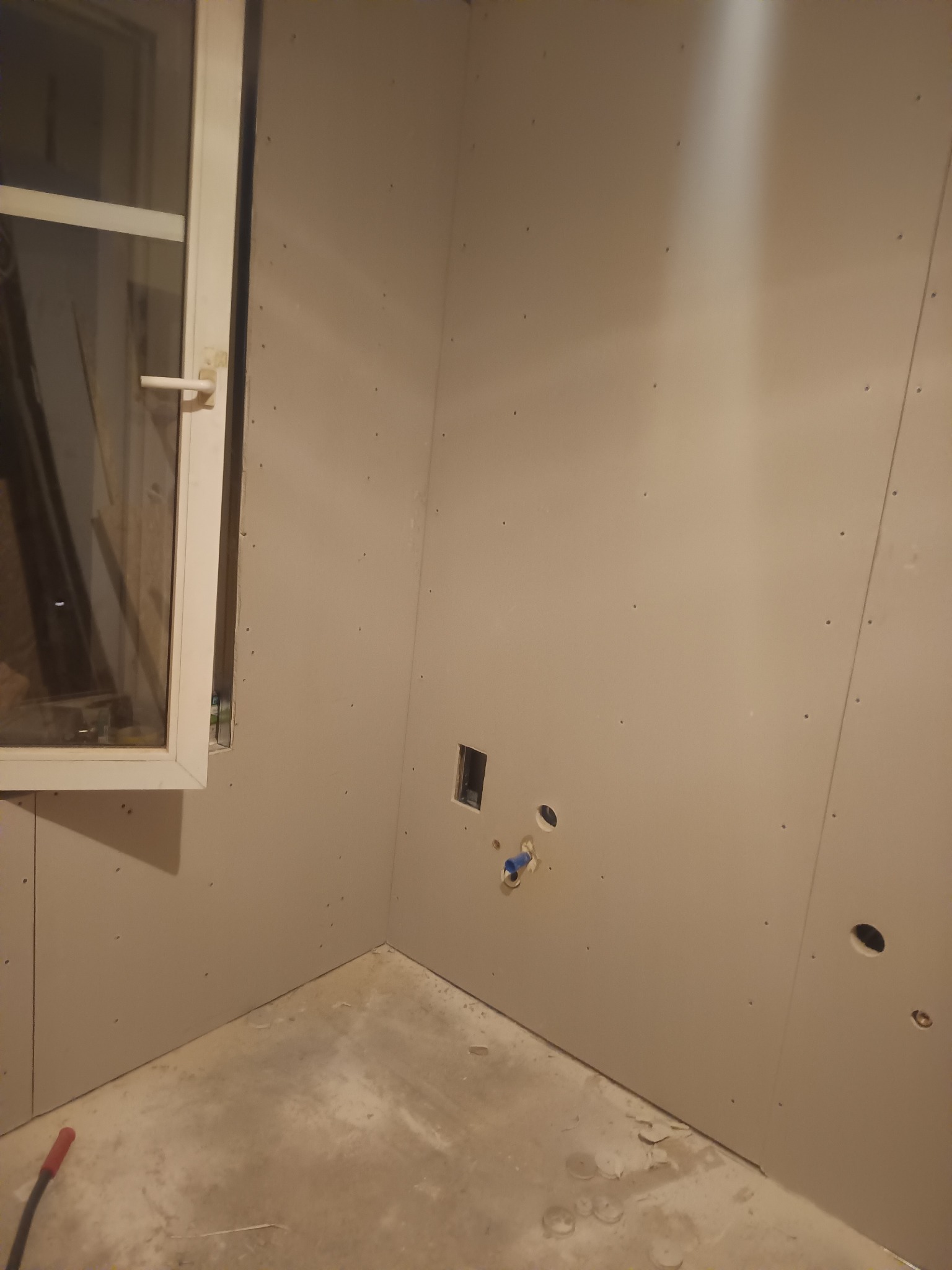 Wnętrze w trakcie prac: ściany pokryte płytami gipsowo-kartonowymi, widoczne otwory na instalacje, surowy beton na podłodze, okno z widocznym zarysem otoczenia.