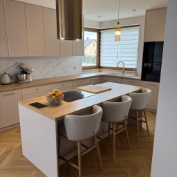 KBmeble - Meble i Kuchnie na wymiar - Jasna kuchnia na wymiar z wyspą, drewnianym blatem i marmurowym backsplashiem. Nad wyspą wisi metalowy okap, a nad blatem złota lampa. Trzy krzesła barowe z jasną tapicerką.