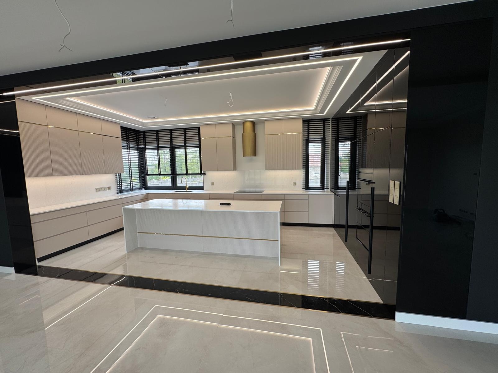 Nowoczesna kuchnia na wymiar z wyspą, beżowe fronty z ryflowaniem, złote akcenty, czarne ramy okienne z żaluzjami i ledowe oświetlenie sufitowe. Wykończenie na wysoki połysk.
