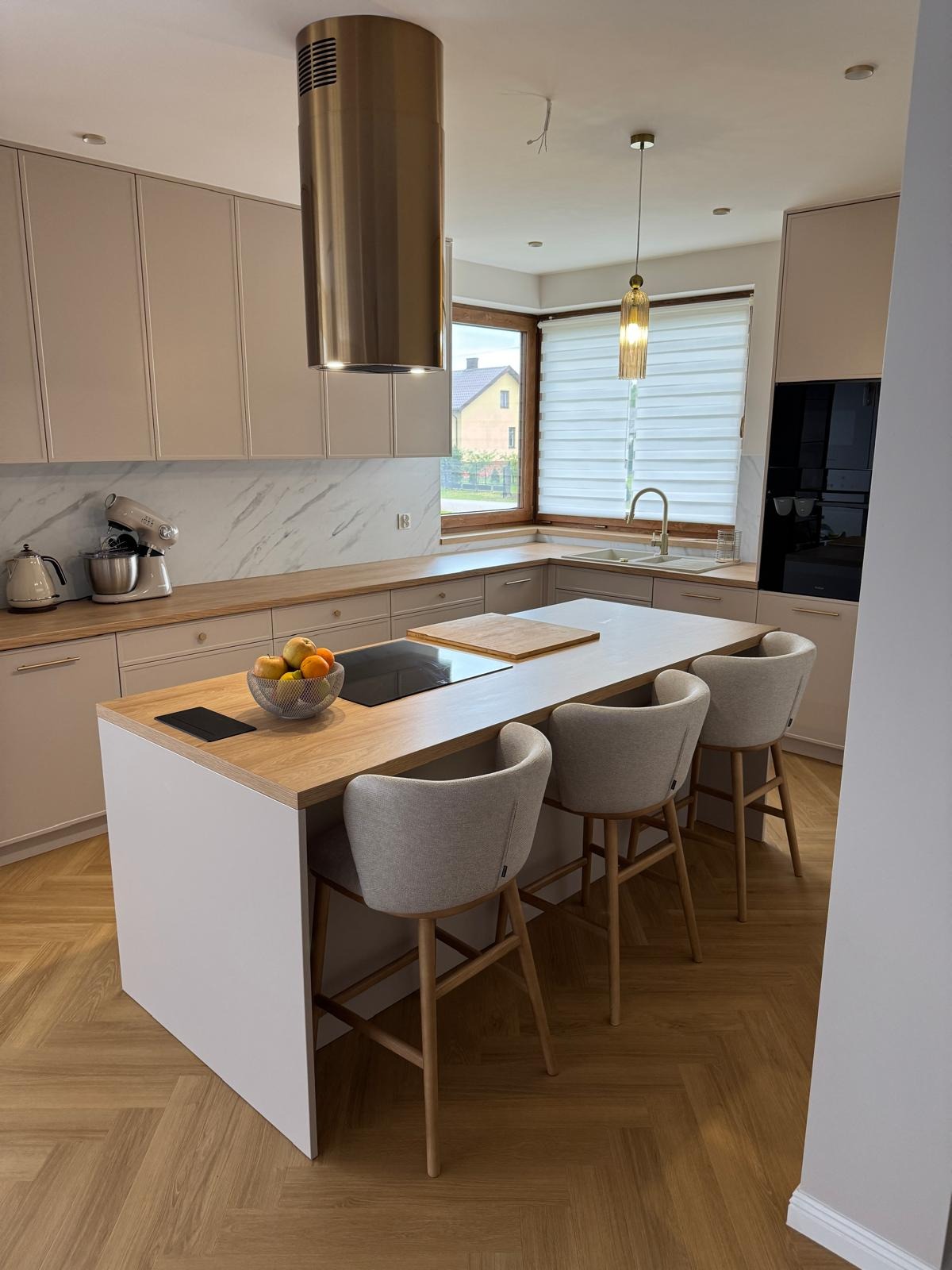Jasna kuchnia na wymiar z wyspą, drewnianym blatem i marmurowym backsplashiem. Nad wyspą wisi metalowy okap, a nad blatem złota lampa. Trzy krzesła barowe z jasną tapicerką.