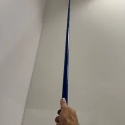 Malowanie ściany wałkiem na długim kiju, widok z dołu, fragment sufitu z oświetleniem. Biała ściana, niebieski kij, ręka malarza w kadrze.