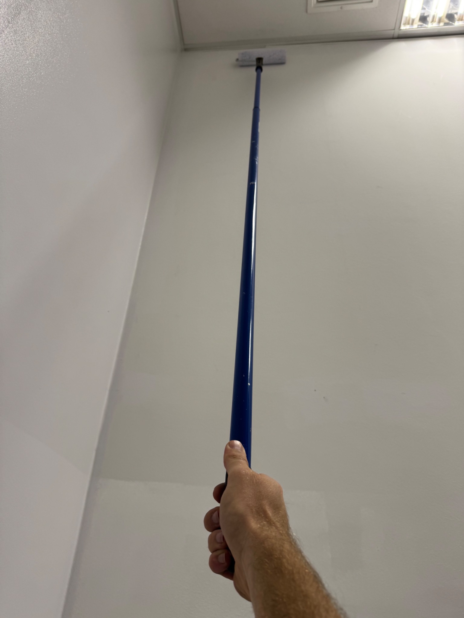 Malowanie ściany wałkiem na długim kiju, widok z dołu, fragment sufitu z oświetleniem. Biała ściana, niebieski kij, ręka malarza w kadrze.
