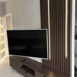 Projektowanie wnętrz Anna Żyjewska-Bas - Nowoczesna szafka RTV z ciemnego drewna, z lameli z oświetleniem LED na ścianie. Minimalistyczny design, jasna podłoga, telewizor na szafce.