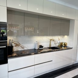 Projektowanie wnętrz Anna Żyjewska-Bas - Nowoczesna kuchnia z białymi frontami i marmurowym blatem, złota armatura, podświetlane szafki górne. Widoczny fragment podłogi w czarno-białe pasy.