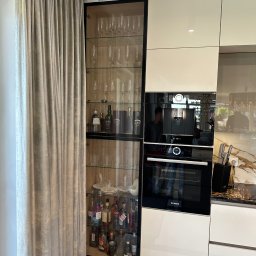 Projektowanie wnętrz Anna Żyjewska-Bas - Biała zabudowa kuchenna z czarną witryną na alkohole i szkło, wbudowany piekarnik i ekspres do kawy. Obok zasłona w odcieniach beżu i szarości.