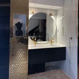 Nowoczesna łazienka z podwójną umywalką, złotymi bateriami i lustrem. Dekoracyjna mozaika i marmurowe płytki dodają elegancji. W tle widoczna rzeźba.
