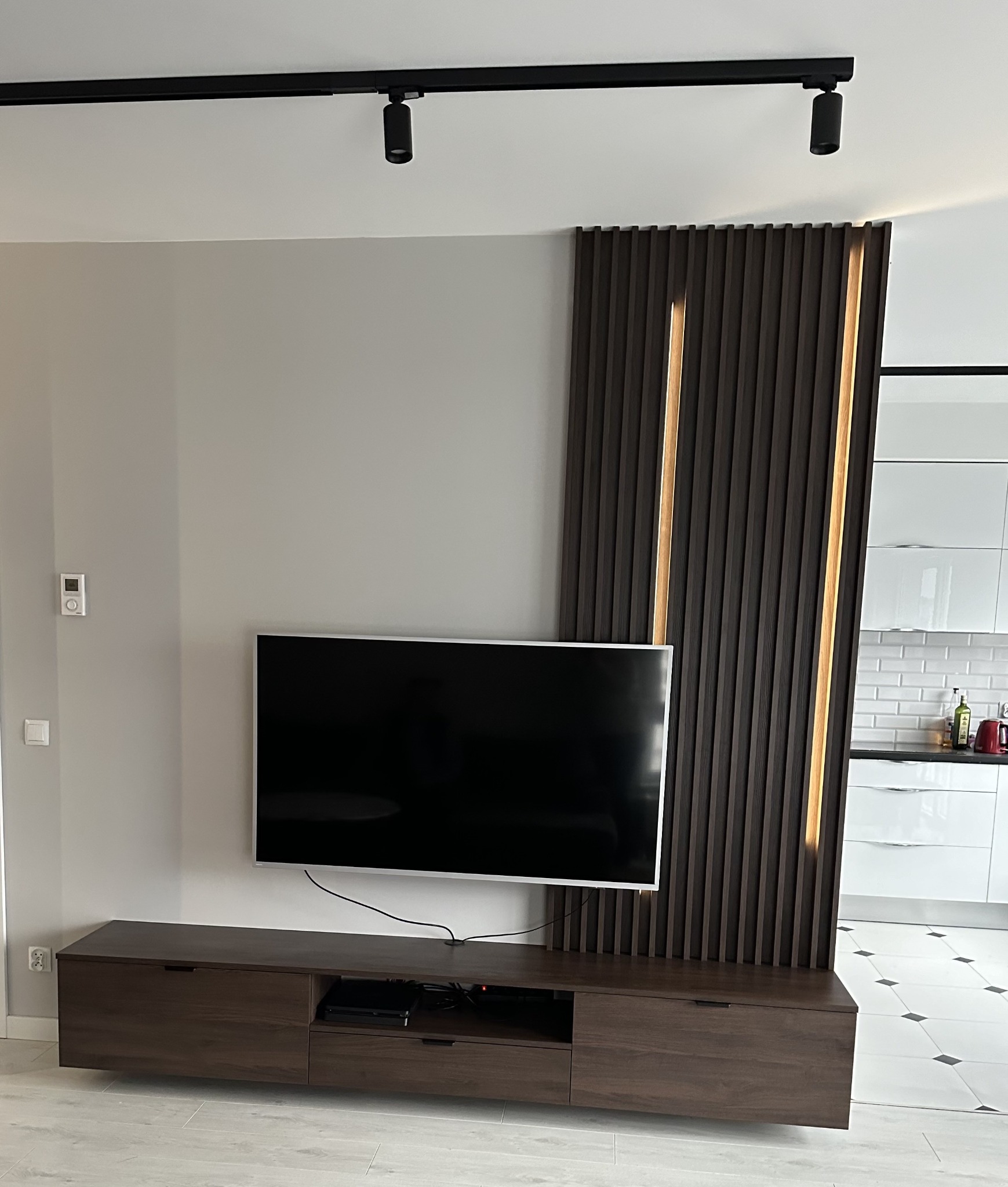 Nowoczesny salon z brązową szafką RTV, telewizorem i dekoracyjną ścianką z lameli z podświetleniem LED. W tle widoczna kuchnia.