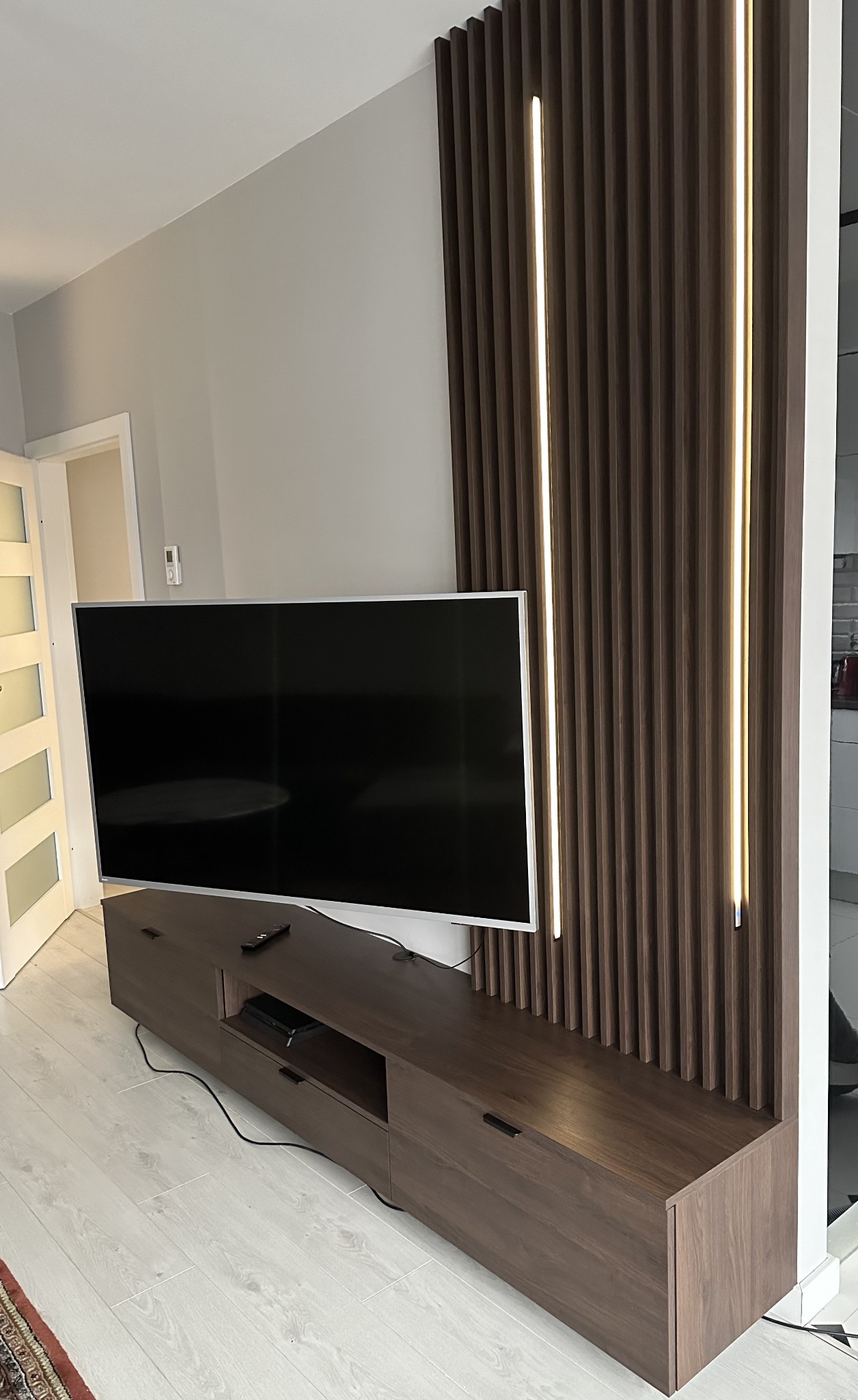 Nowoczesna szafka RTV z ciemnego drewna, z lameli z oświetleniem LED na ścianie. Minimalistyczny design, jasna podłoga, telewizor na szafce.