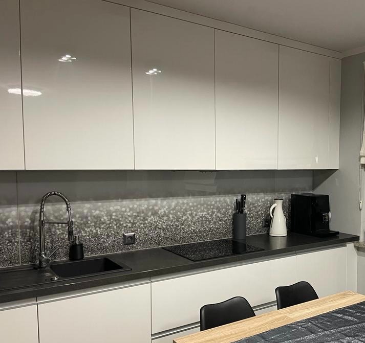 Biała, nowoczesna kuchnia z błyszczącymi szafkami, czarnym blatem i dekoracyjnym panelem nad blatem. Widoczny zlew, płyta indukcyjna i ekspres do kawy.