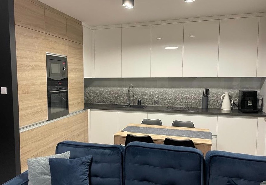 Nowoczesna kuchnia z zabudową w odcieniach bieli i drewna, zintegrowany piekarnik i mikrofalówka, stół z krzesłami, sofa w kolorze granatowym.