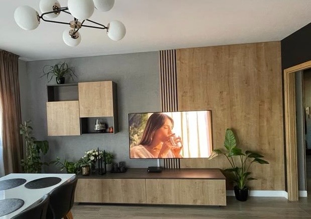 Nowoczesny salon z drewnianą ścianą, podświetlanym telewizorem, wiszącymi szafkami i roślinami. Minimalistyczny design, ciepłe oświetlenie, widok na jadalnię.