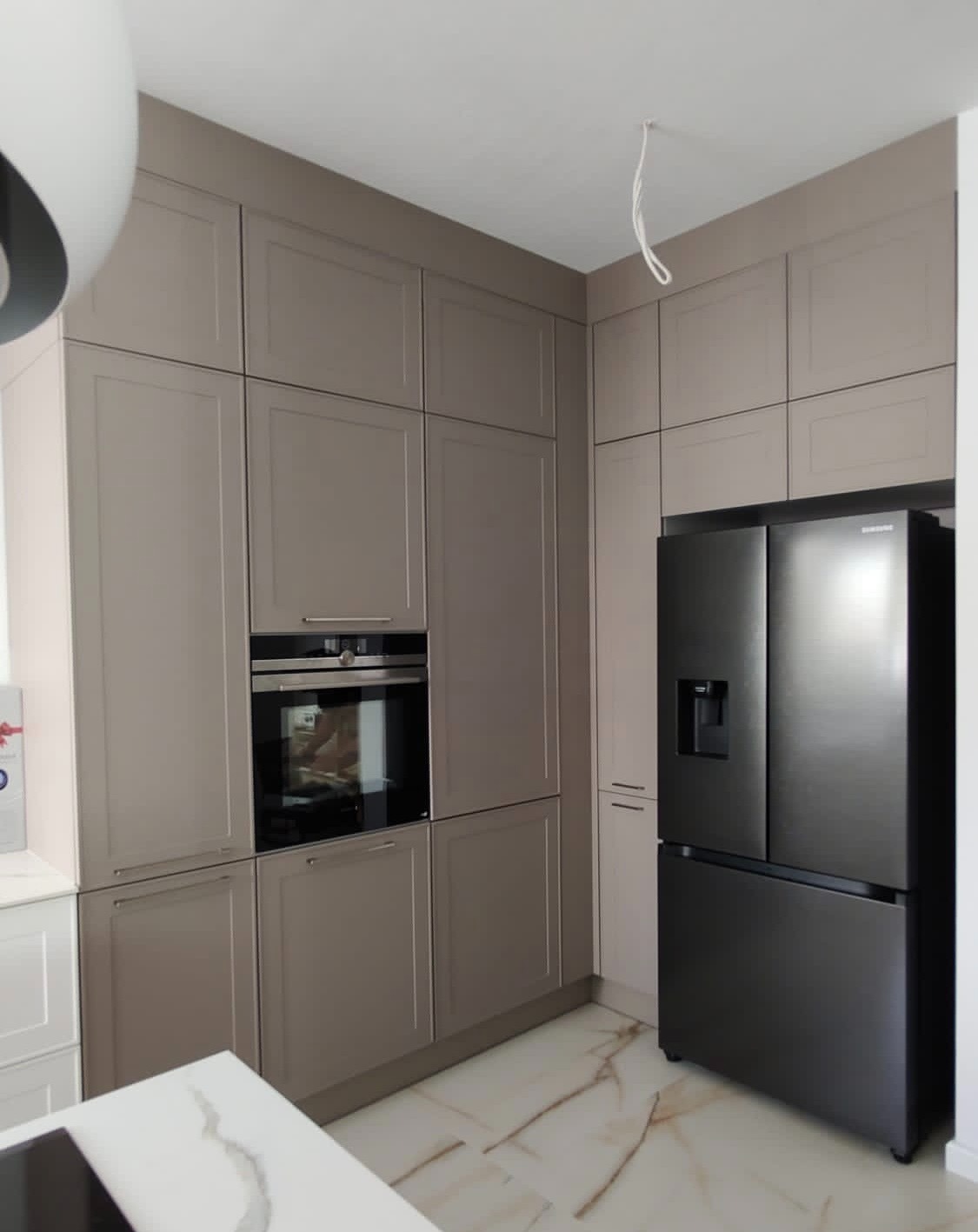 Nowoczesna kuchnia w odcieniach beżu z zabudową na wymiar, piekarnikiem i czarną lodówką typu side-by-side. Marmurowe płytki podłogowe.