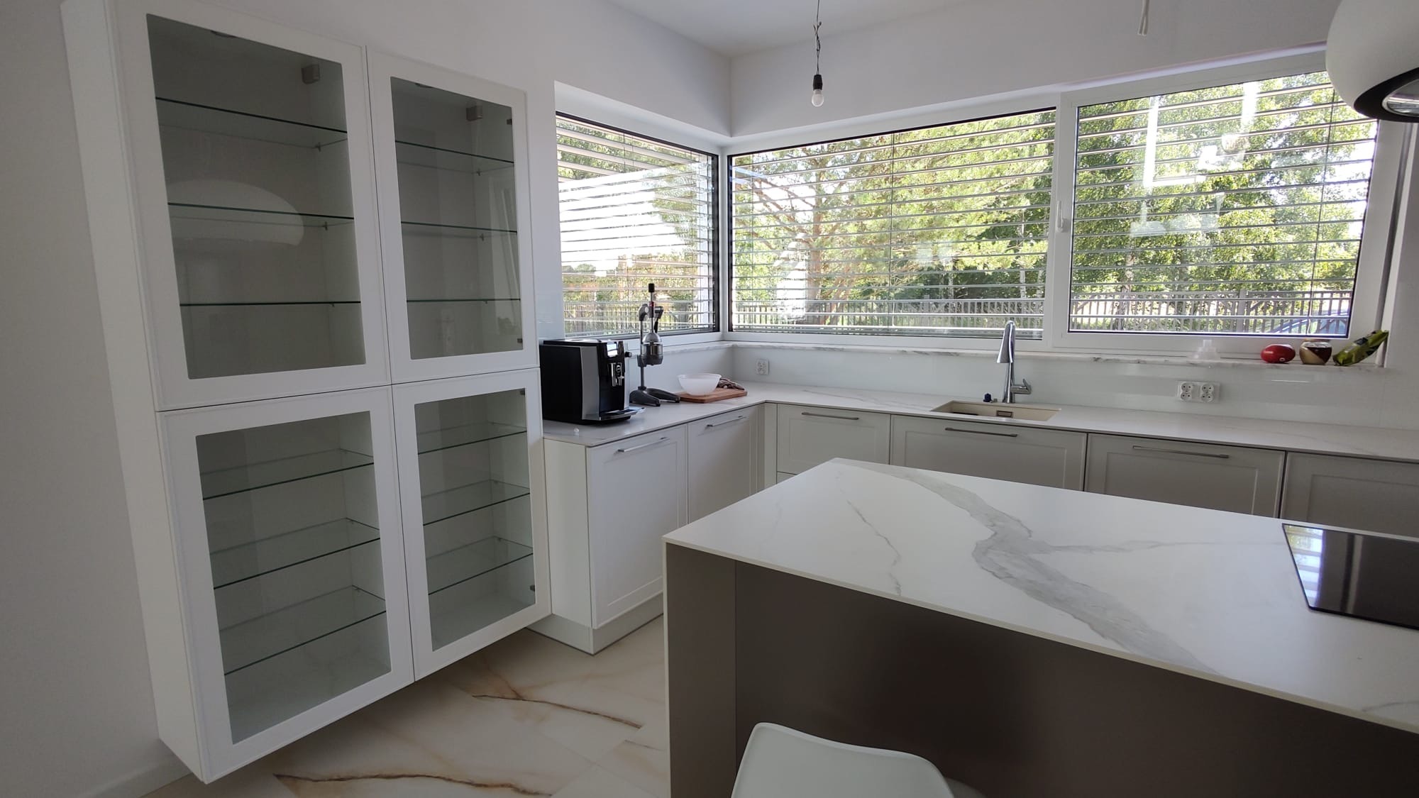 Nowoczesna kuchnia z wyspą i dużym oknem z widokiem na zieleń. Białe szafki z przeszkleniami, marmurowy blat i subtelne oświetlenie tworzą elegancką przestrzeń.