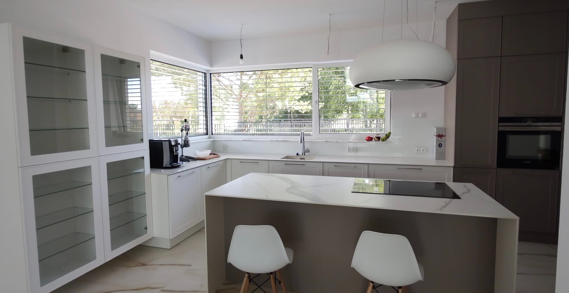 Jasna kuchnia z wyspą i oknem z widokiem na zieleń. Fronty szafek białe i szare, blaty z marmurowym wzorem. Nad wyspą okrągła lampa. Na blacie ekspres do kawy.