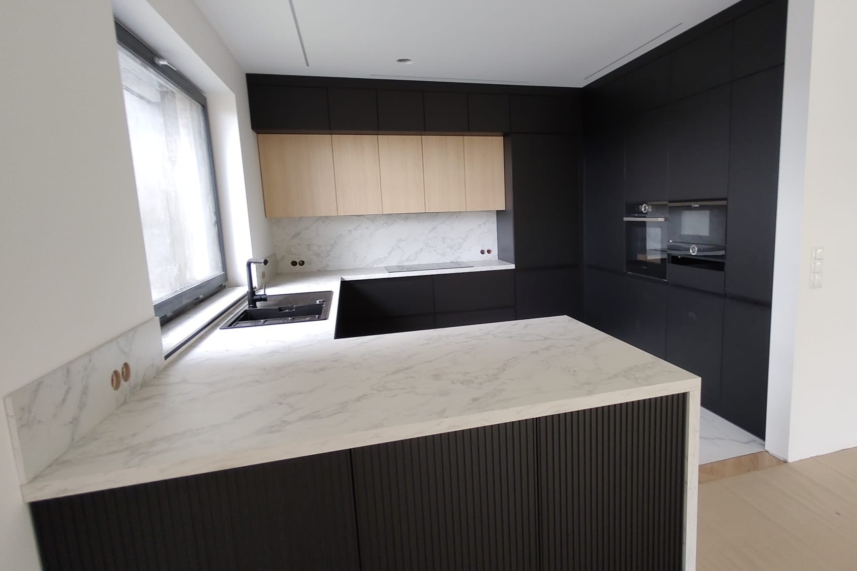 Nowoczesna kuchnia z wyspą: blaty marmurowe, czarne szafki z ryflowanym frontem wyspy, drewniane górne szafki. Minimalistyczny design, jasne wnętrze.