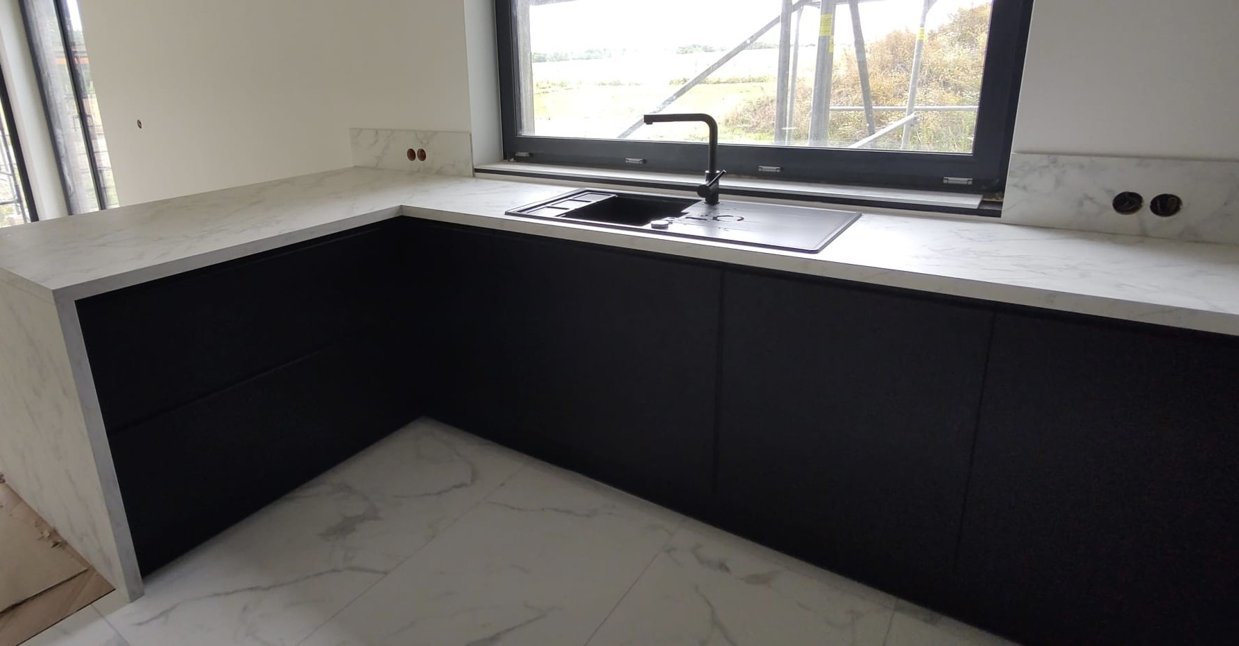 Nowoczesna, czarna zabudowa kuchenna z blatem imitującym marmur i widokiem na zewnątrz przez okno. Elementy wykończeniowe z marmuru. Minimalistyczny design.