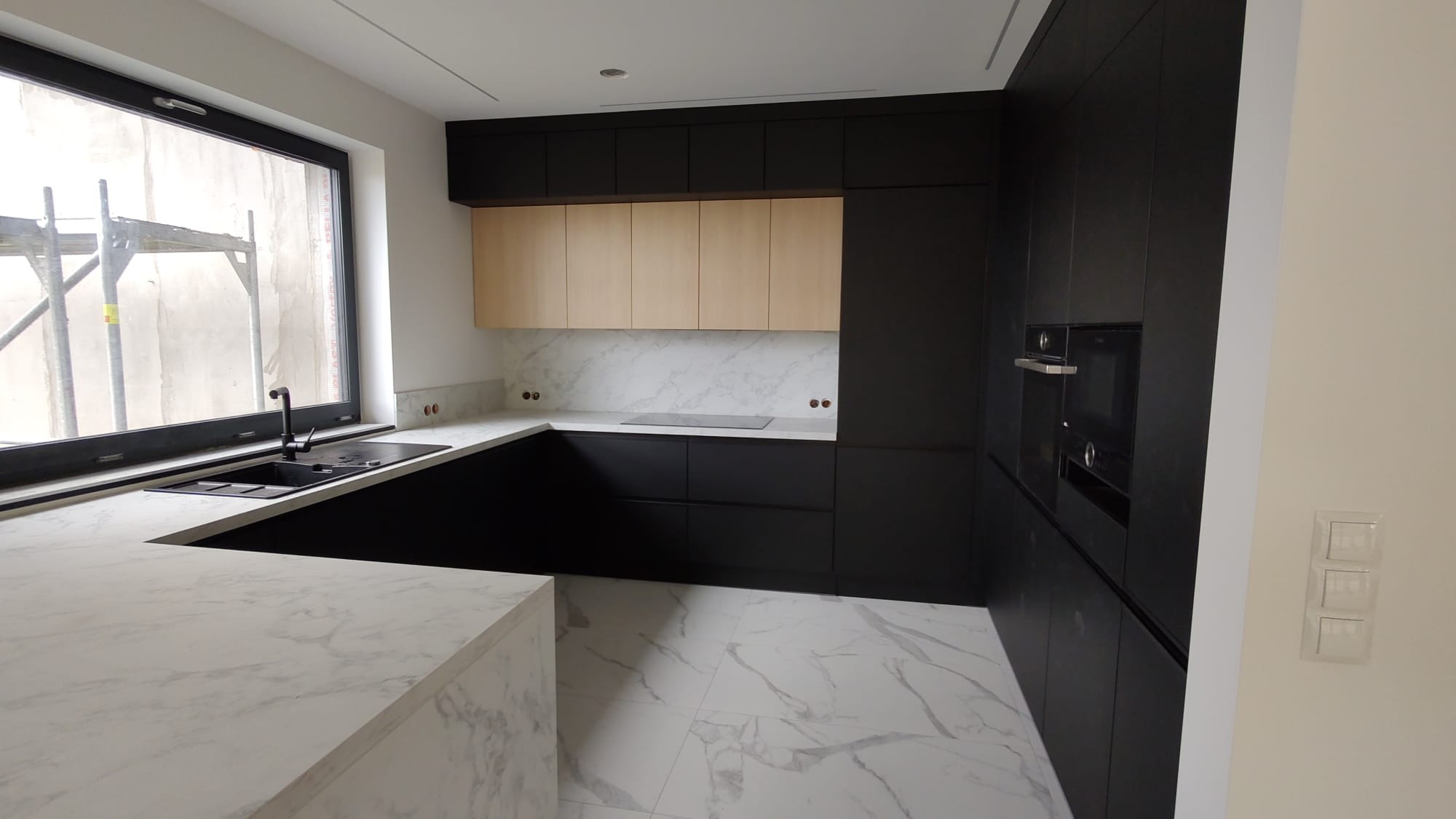 Nowoczesna, czarno-beżowa kuchnia z marmurowym blatem i podłogą. Widok na okno z rusztowaniem na zewnątrz. Minimalistyczny design, wbudowane sprzęty.