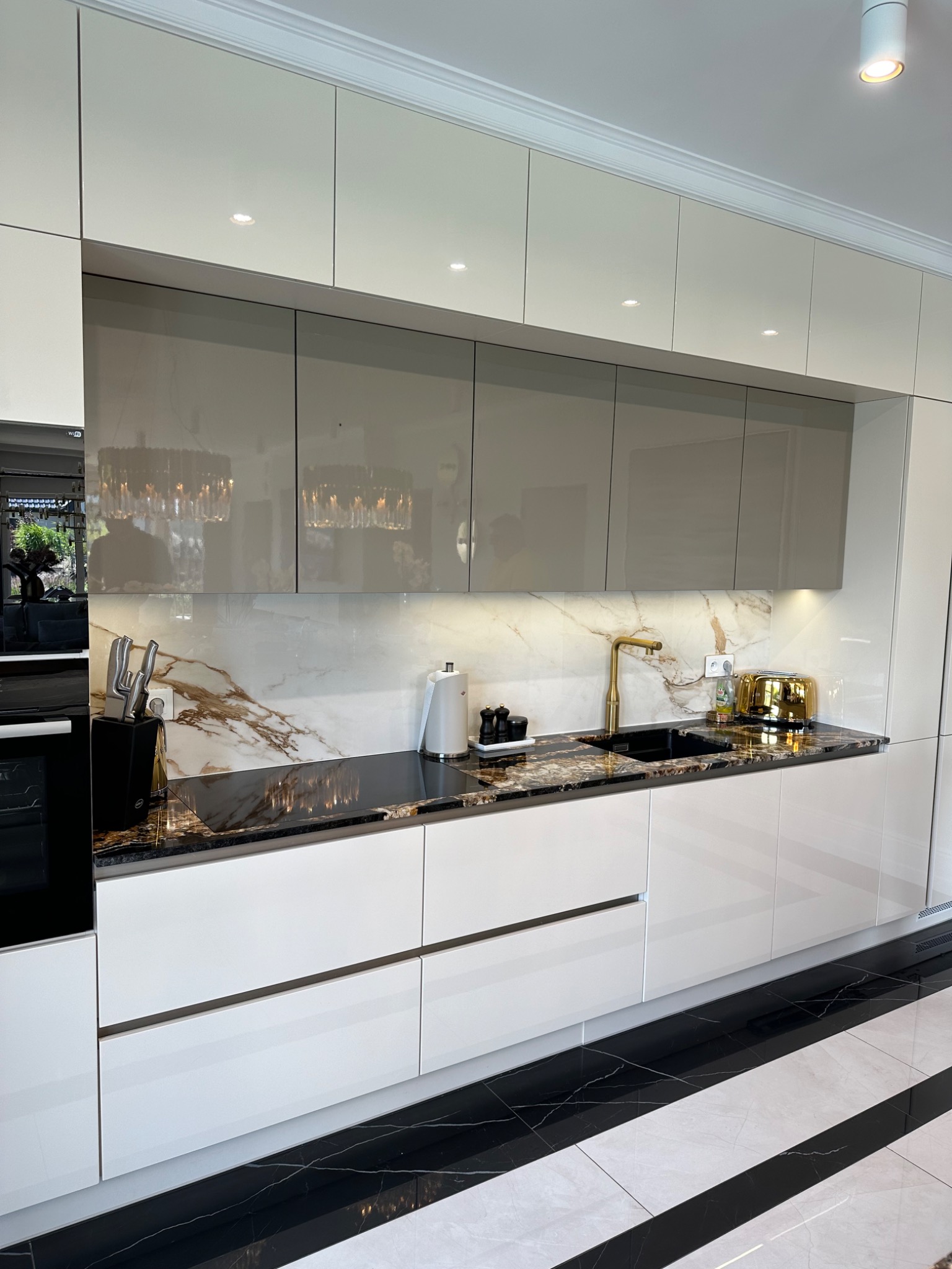 Nowoczesna kuchnia z białymi frontami i marmurowym blatem, złota armatura, podświetlane szafki górne. Widoczny fragment podłogi w czarno-białe pasy.