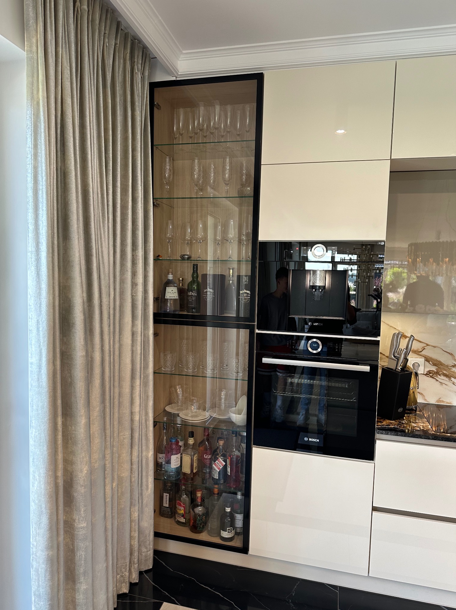 Biała zabudowa kuchenna z czarną witryną na alkohole i szkło, wbudowany piekarnik i ekspres do kawy. Obok zasłona w odcieniach beżu i szarości.