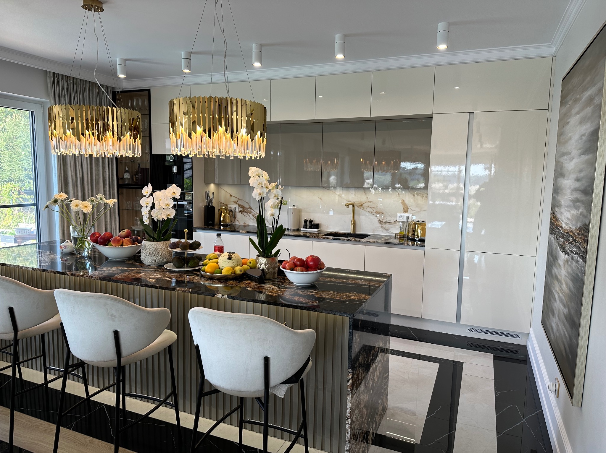 Elegancka kuchnia z wyspą, oświetlona złotymi lampami. Marmurowy blat, białe szafki i krzesła barowe tworzą luksusową przestrzeń. Na wyspie owoce i kwiaty.