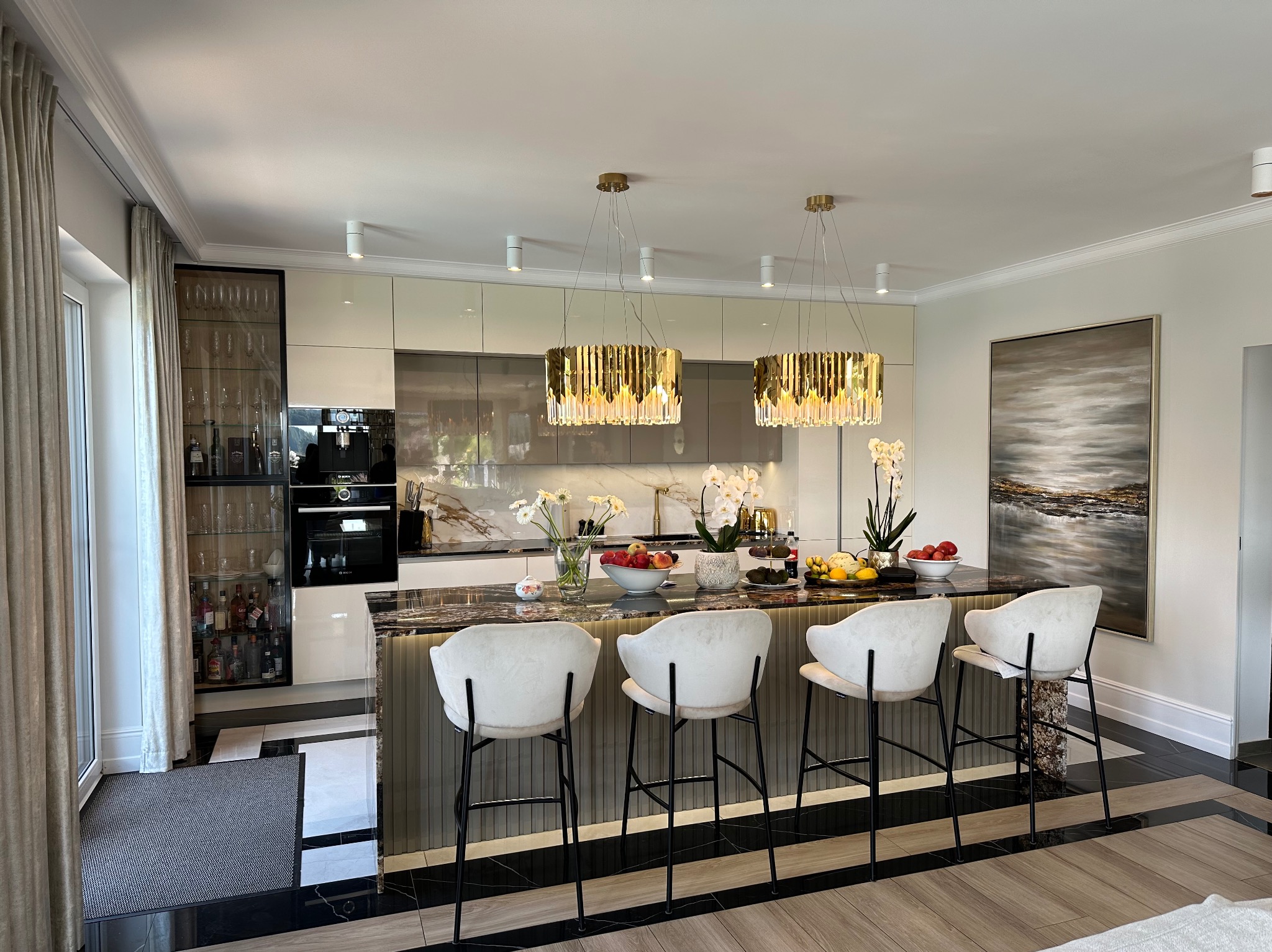 Elegancka kuchnia z wyspą i czterema krzesłami barowymi. Marmurowy blat, złote lampy wiszące, przeszklona witryna i nowoczesne szafki w beżowej tonacji.