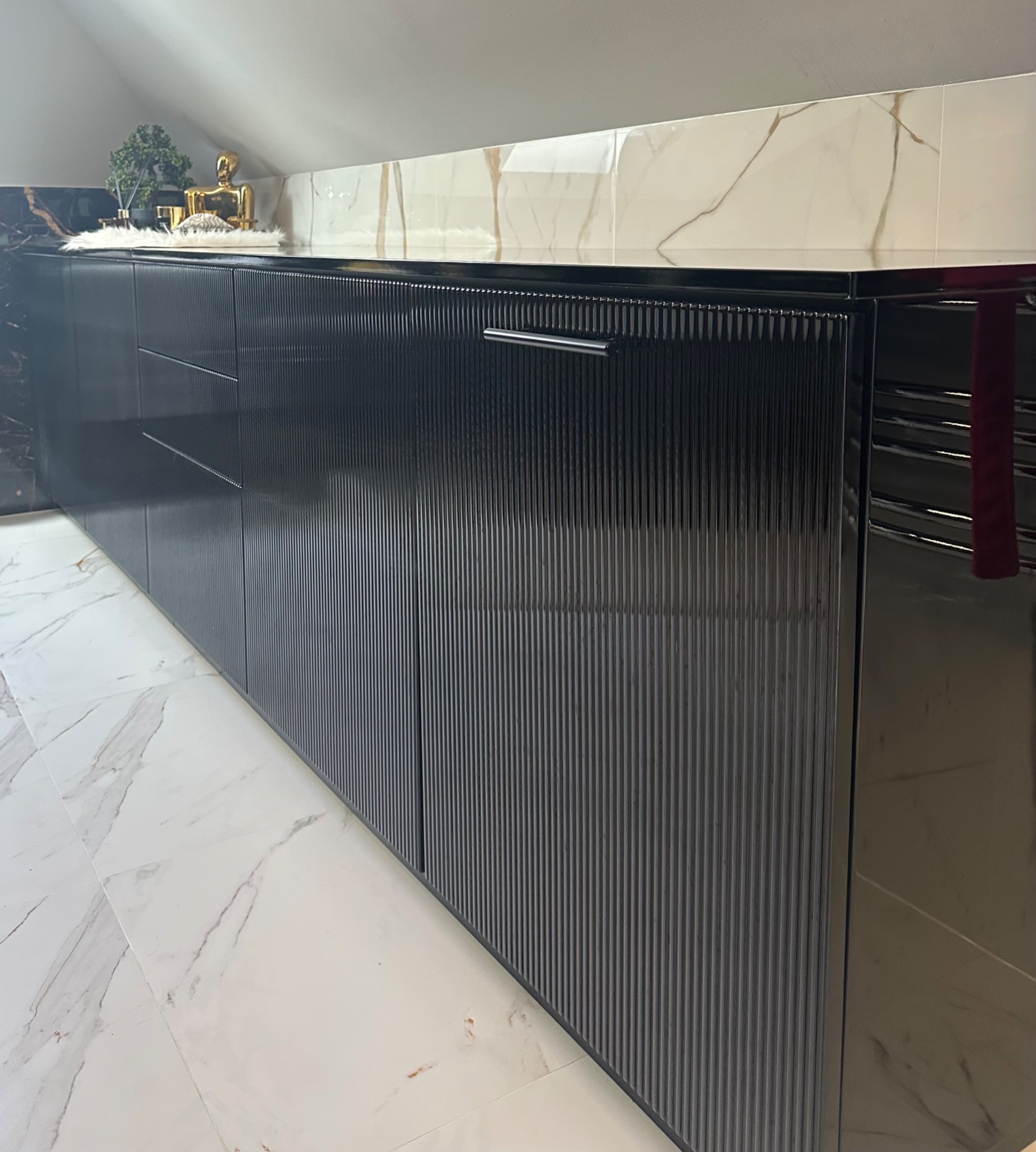 Czarne, lakierowane szafki kuchenne z ryflowanymi frontami, wykończone blatem z marmuru. Na blacie dekoracje: figurka i roślina. Podłoga z płytek imitujących marmur.